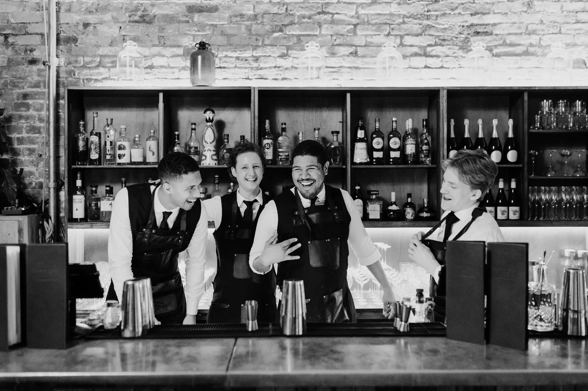 Four smiling bartenders in aprons behind a bar