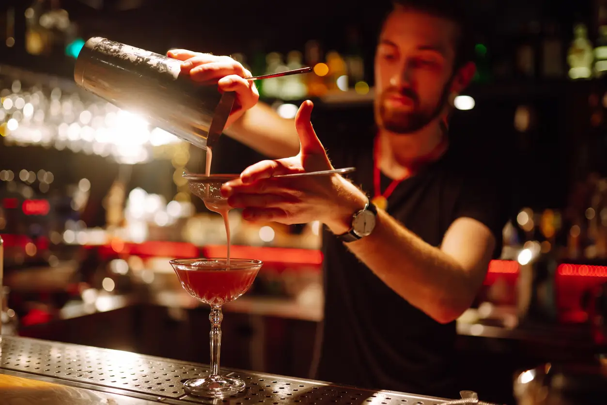 A bartender pouring a red cocktail from a shaker into a coupe glass